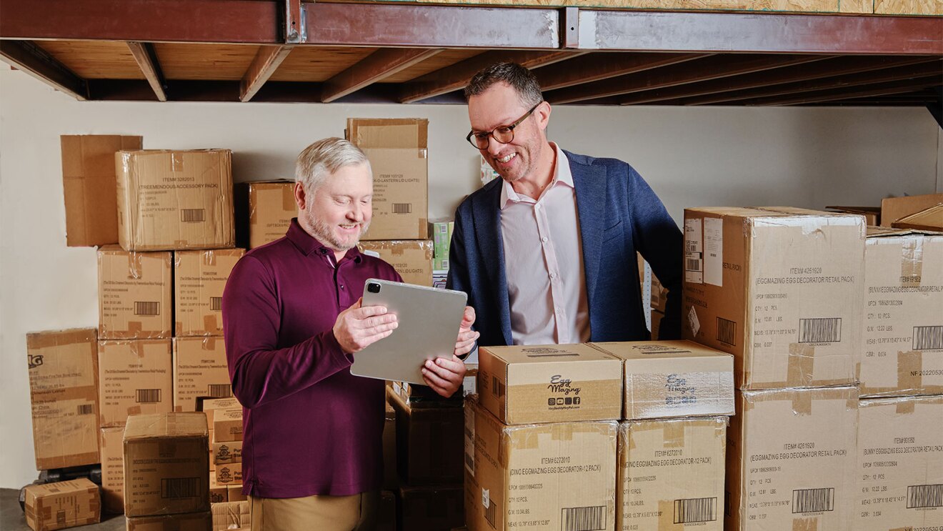 Two businessmen reviewing inventory on tablet in warehouse full of boxes