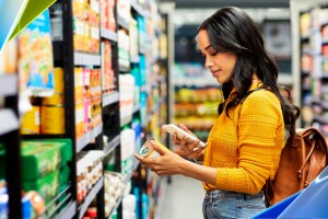 A woman stands in a grocery store aisle and holds a can of food while looking at her phone.