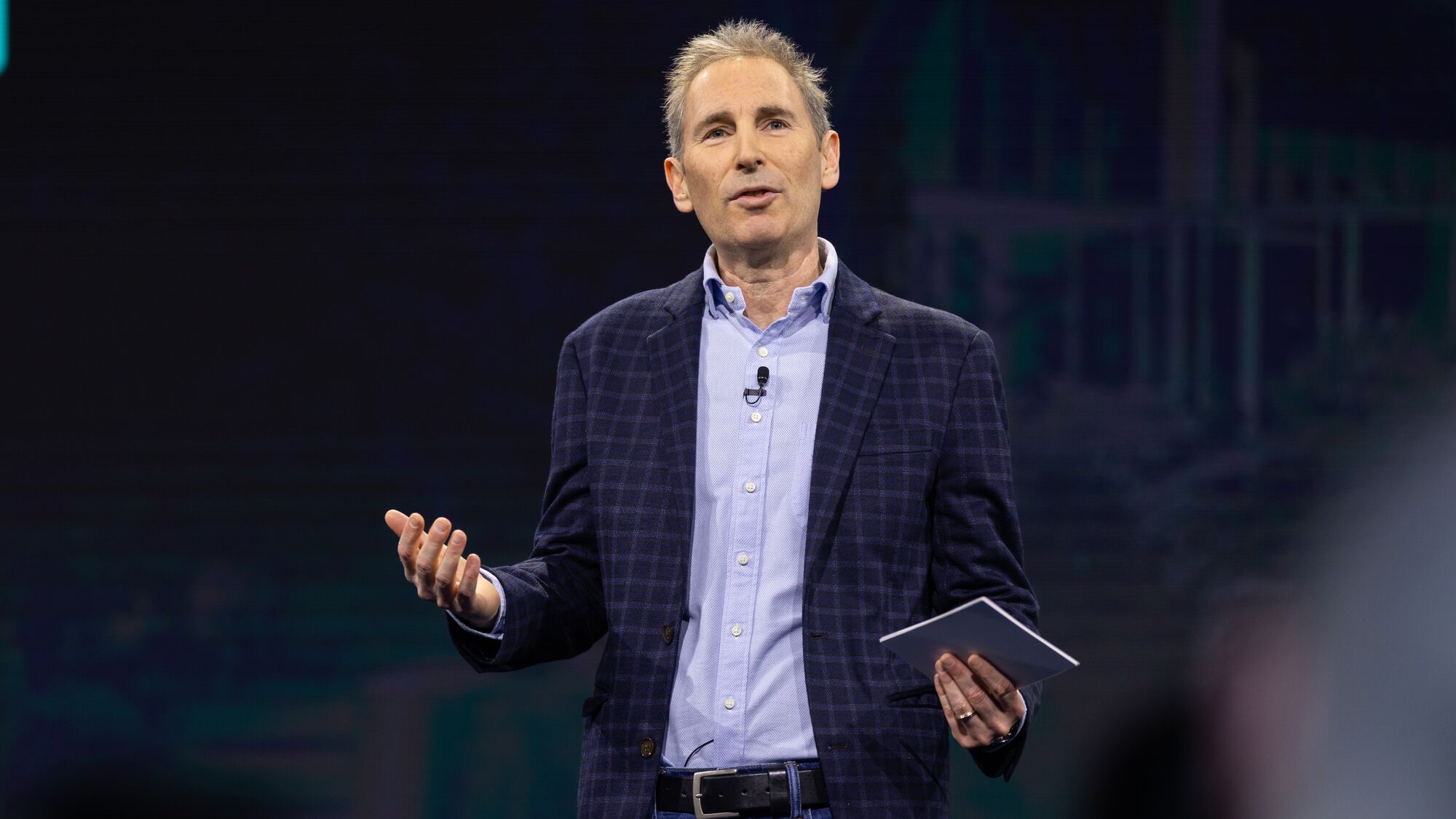 Andy Jassy wearing a plaid blazer presenting on large stage in front of Amazon employees.